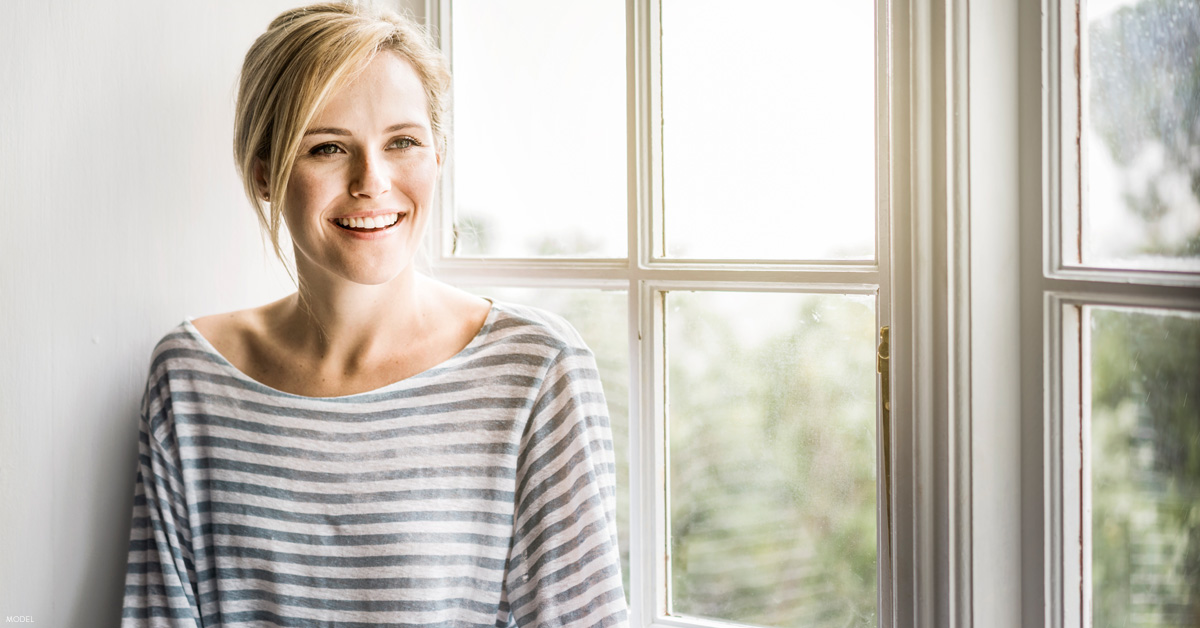 Woman standing by window in St. Louis considering tummy tuck vs. liposuction procedures